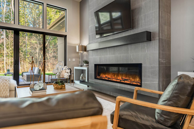 Modern living room with a linear electric fireplace built into a gray tile wall beneath a wall-mounted TV, with floor-to-ceiling windows overlooking trees