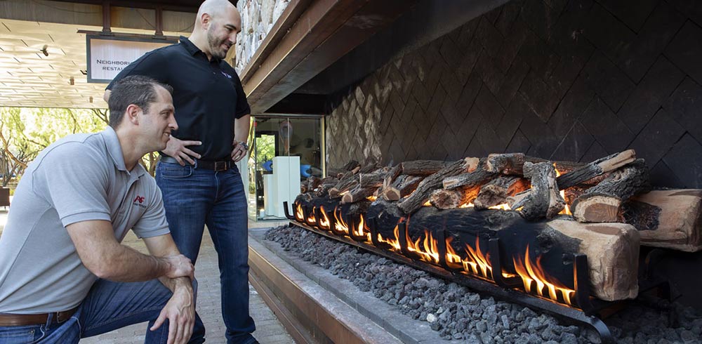 Two installers inspecting a lit Grand Canyon gas log set in an outdoor stone fireplace